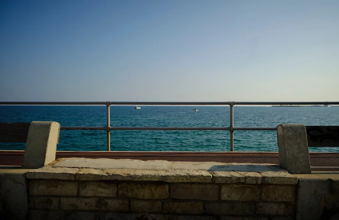 Stone bench by a seaside railing in Colònia de Sant Jordi, overlooking calm blue water and a clear sky with distant boats—perfect for enjoying beachfront ceviche.