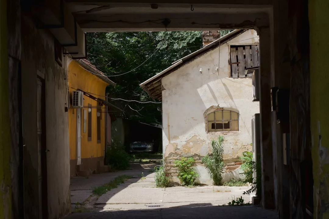 A narrow alley with old yellow and white buildings, overgrown plants, and a car parked in the shade evokes the charm of Santa Eugenia—perfect for those considering buying a renovated farmhouse nearby.