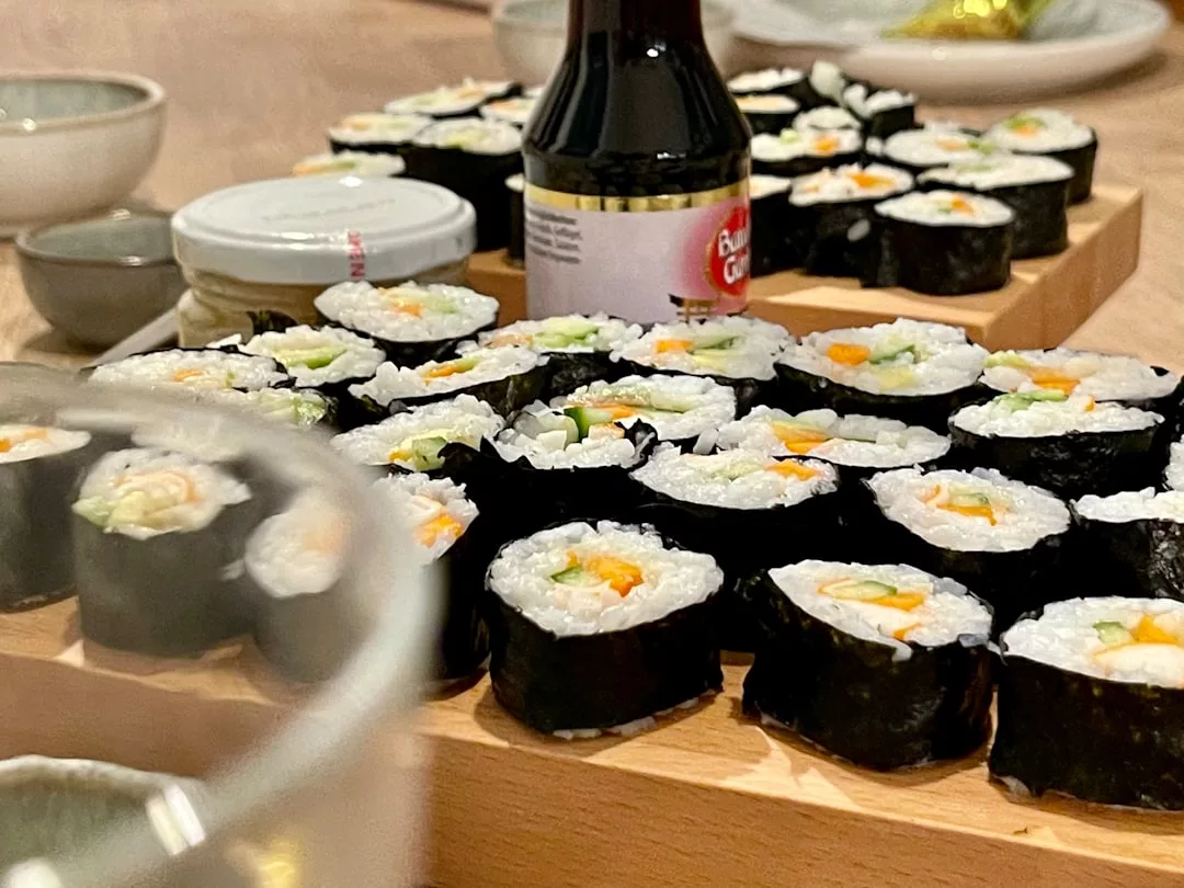 Close-up of assorted sushi rolls with rice, vegetables, and nori on a wooden board, created during chef-led sushi workshops in Port de Sóller, with soy sauce in the background.