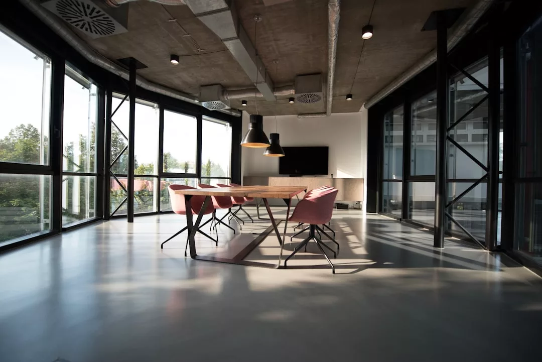Modern conference room with large windows, a long table, pink chairs, and pendant lights—ideal for commercial office spaces for rent in Palma Nova.