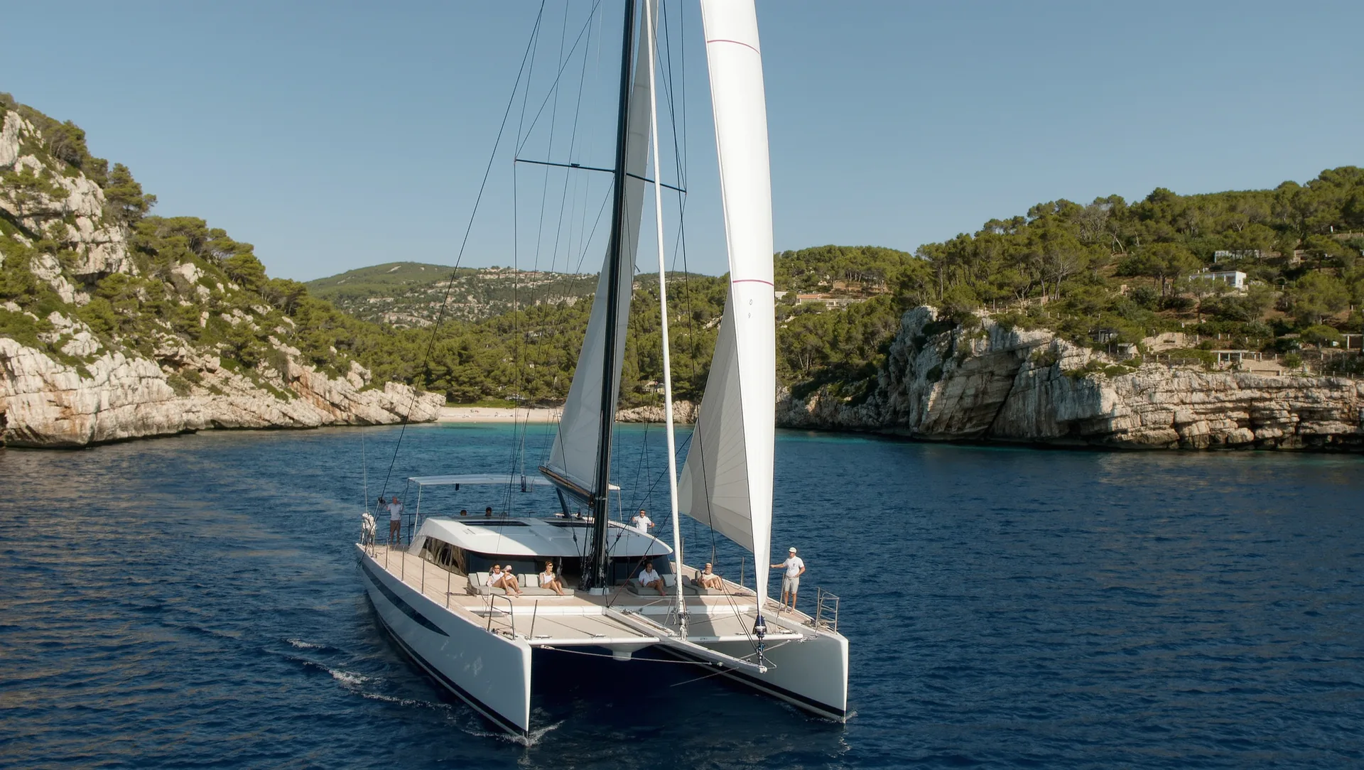 A crewed sailing catamaran under full sail off the south coast of Mallorca, crew trimming the mainsail and guests on the forward deck.
