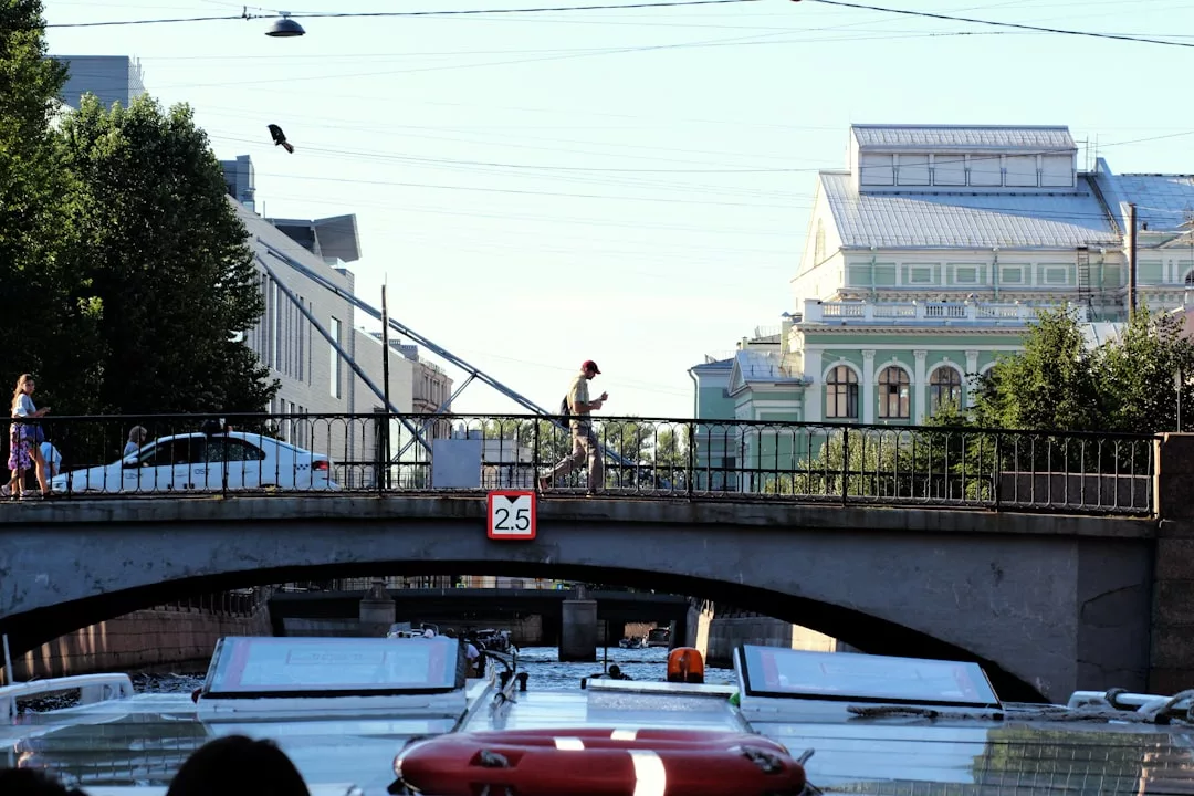 A person walks across a bridge over a canal in Sant Elm, with buildings, trees, and hints of boat trips to Dragonera Island in the background.