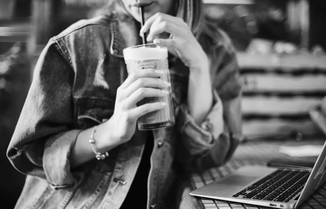 Woman in a denim jacket drinking an iced coffee with a straw, sitting by an open laptop, browsing winery-restaurants in Biniagual and exploring tasting rooms for her next weekend getaway.