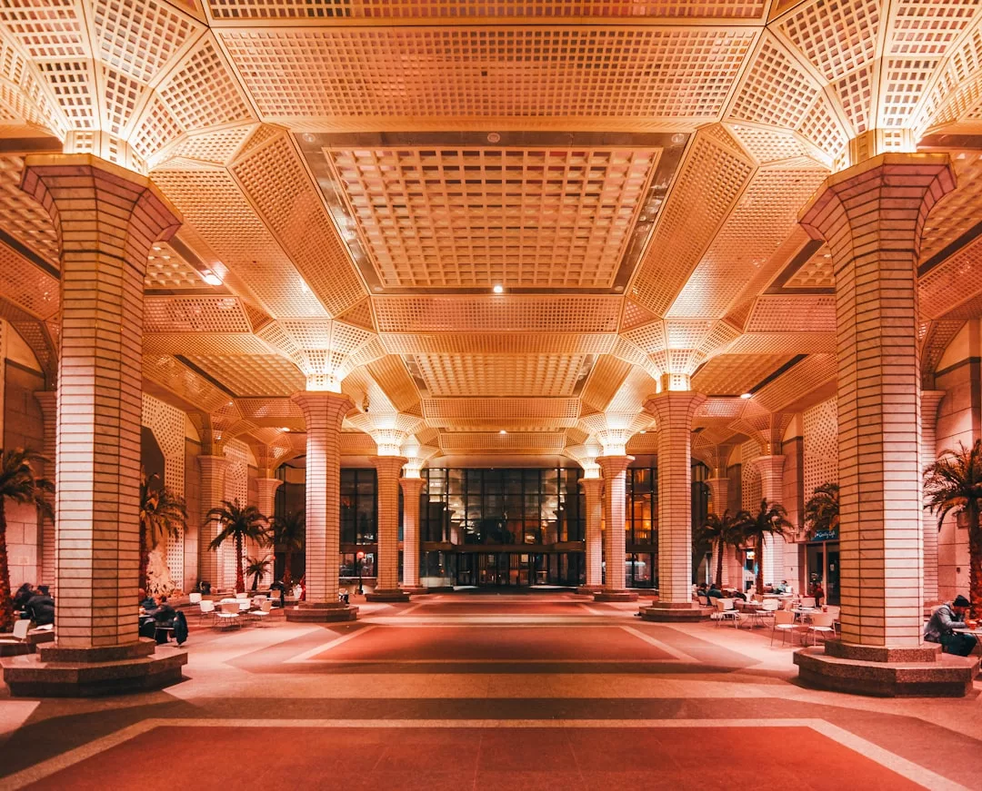 Indoor hall with large columns, patterned ceiling, and palm trees evokes the elegance of town palaces in Palma, with people sitting on benches under warm lighting—an ambiance reminiscent of historic accommodations.