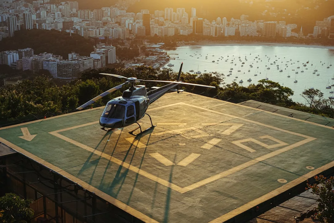 A helicopter on a rooftop helipad overlooks a coastal city and bay at sunset, offering the perfect arrival for guests enjoying VIP Helicopter Transfers to luxurious Mallorca Resorts.