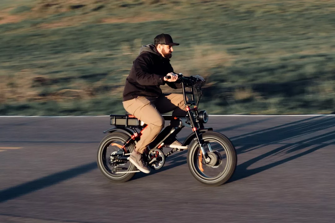 Man riding a black electric bike on a paved road near Valldemossa, with olive groves and grassy background, wearing a jacket and cap.