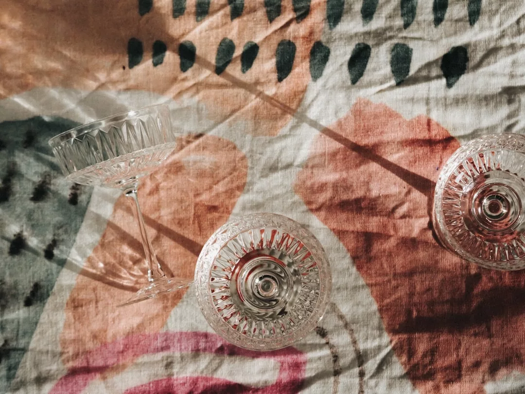 Two crystal glasses casting shadows on a colorful, patterned fabric in sunlight, evoking the magic of fairy-tale ceremonies in Alcúdia’s elegant wedding suites.