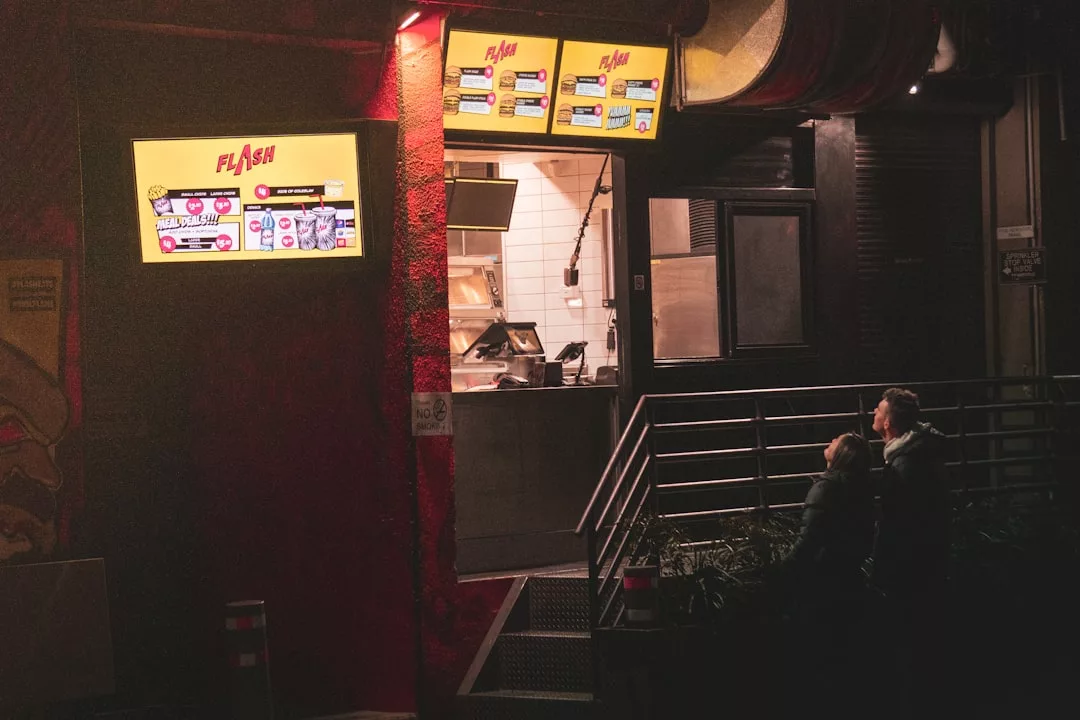 Two people sit outside a fast food restaurant at night on the Santa Ponsa beachfront promenade, looking up at the brightly lit menu boards.