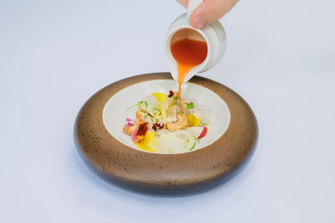 A hand pours orange sauce onto a plated gourmet dish with vegetables, flowers, and garnishes—an artful creation inspired by gourmet restaurants in Sóller, Mallorca.