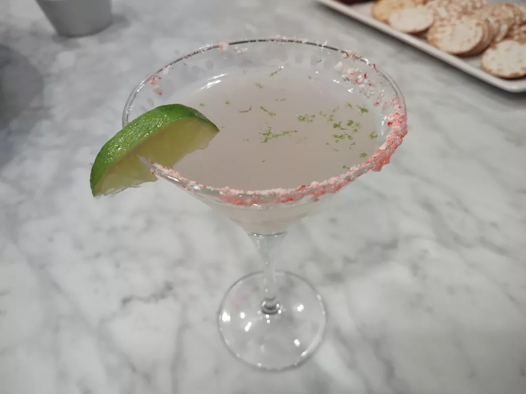 A cocktail in a martini glass with a lime wedge and pink sugar rim, served on a marble table at one of Magaluf’s top cocktail restaurants, crackers in the background.