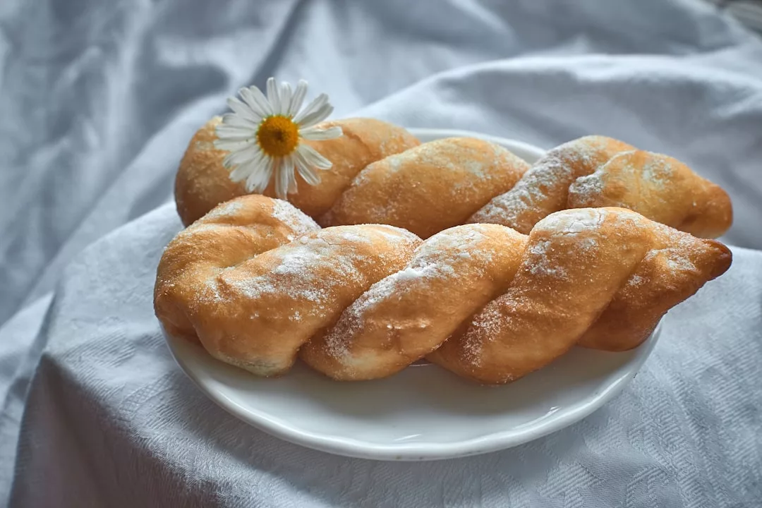 Twisted sugar-dusted pastries on a white plate with a daisy flower on top, set on a white fabric background—just like the treats in Palma’s famed dessert parlors near La Lonja.