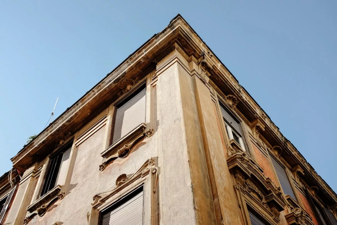 Upward view of an old building corner in Valldemossa, its closed shutters and ornate details reflecting authenticity and historic property preservation against a clear sky.