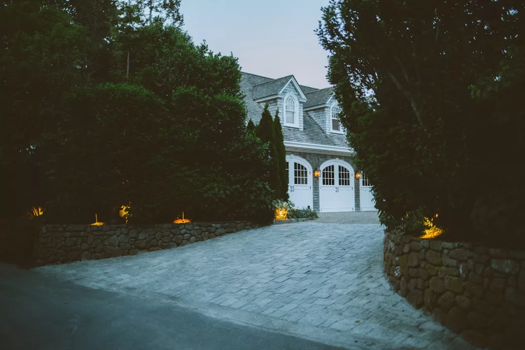 A luxurious large house with three white garage doors and an illuminated stone driveway, perfect for romantic dinners, surrounded by trees at dusk—reminiscent of enchanting Deià evenings.