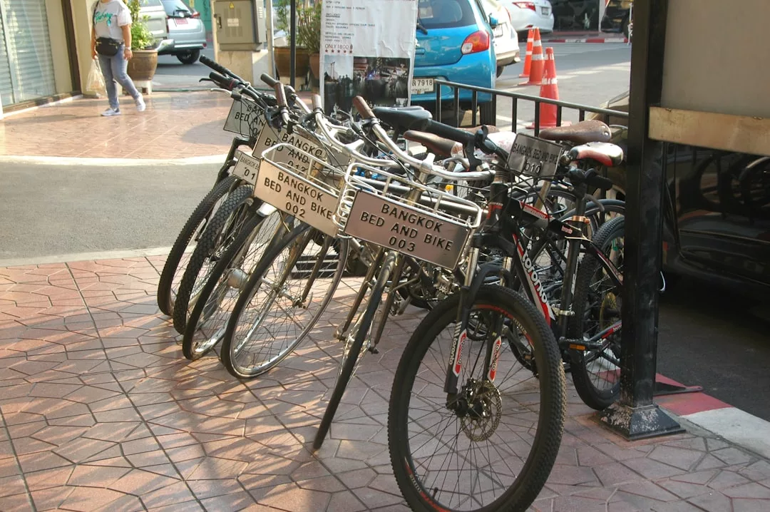 Several bicycles with "Bangkok Bed and Bike" signs, including a few luxury e-bike rental options, are parked on a tiled sidewalk near a street.