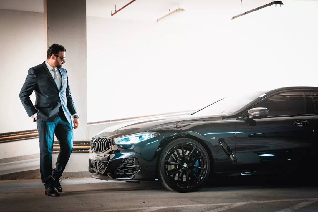 Man in suit stands next to a sleek black car in a brightly lit indoor parking garage, capturing the upscale vibe of Palma’s Santa Catalina district, known for its luxury gastro-pubs.