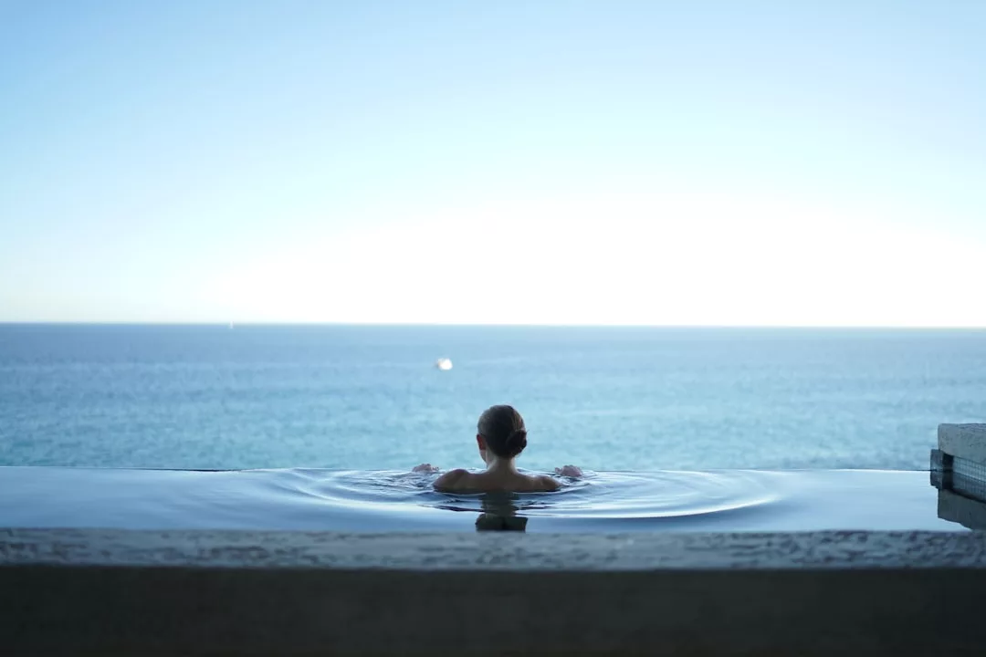 Person relaxing in an infinity pool at one of the luxury hotels Deià, overlooking a calm, blue ocean under a clear sky with the Tramuntana mountains in the distance.
