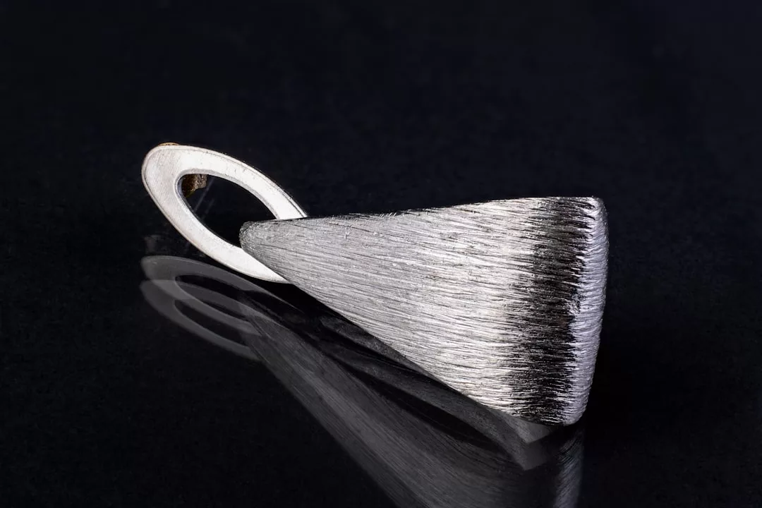 A silver, triangular pendant with a textured surface lies on a dark, reflective background, evoking the elegance of luxury photoshoots or the timeless charm of Mediterranean backdrops.