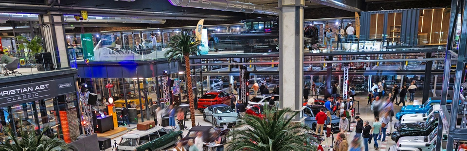 Large indoor car showroom in Mallorca featuring classic and modern luxury vehicles, glass-walled offices above, and groups of people walking and observing the cars.