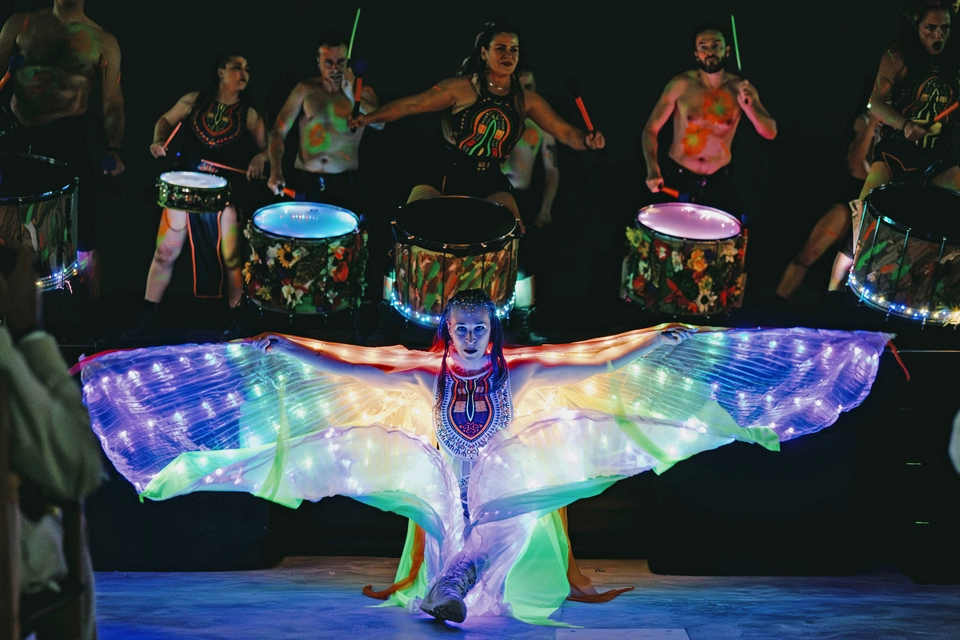 A performer with illuminated wings poses in front of drummers with painted bodies and neon drumsticks on a dark stage, creating a scene of luxury entertainment inspired by Mallorca’s vibrant nightlife.