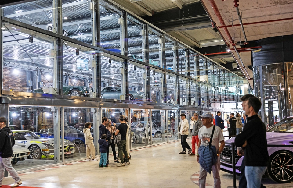 People gather inside a modern luxury car showroom in Mallorca, featuring two levels of glass-enclosed parking displaying various high-end cars.