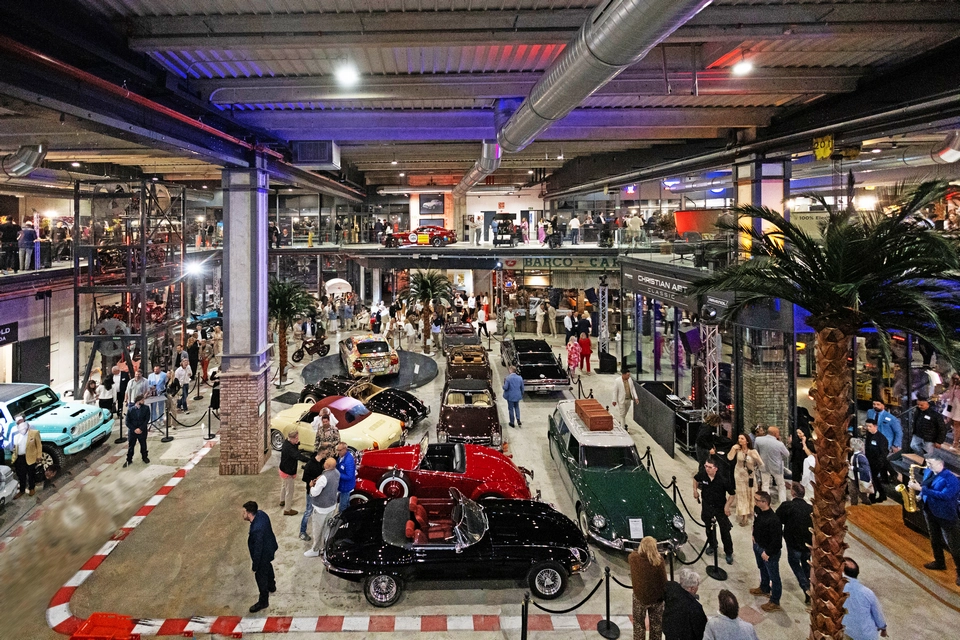 Indoor car exhibition featuring luxury classic cars on display, surrounded by crowds of people under bright lights in a spacious, industrial-style venue reminiscent of Mallorca’s vibrant event scene.