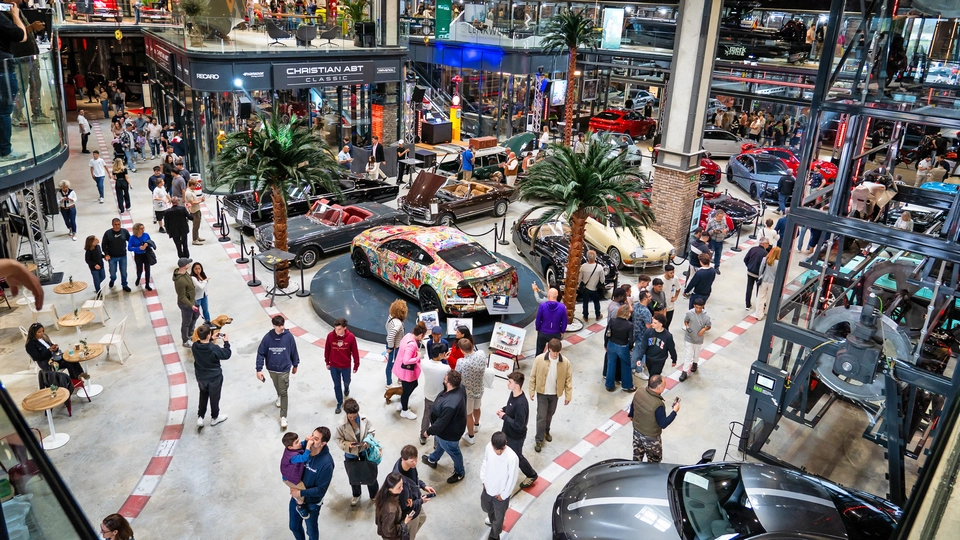 A busy indoor car exhibition features people walking among various classic and luxury modern cars, with palm trees and seating areas scattered throughout the Mallorca-inspired venue.