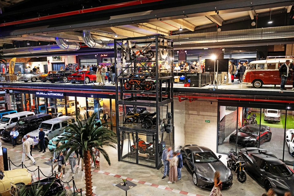 Indoor car showroom in Mallorca featuring luxury classic and modern vehicles on display, a central motorcycle elevator, and people walking around and viewing the exhibits.