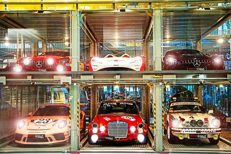 Six classic and modern luxury sports cars are displayed in a two-level glass car storage facility, with three cars on each level, under bright lighting—evoking the elegance of Mallorca.