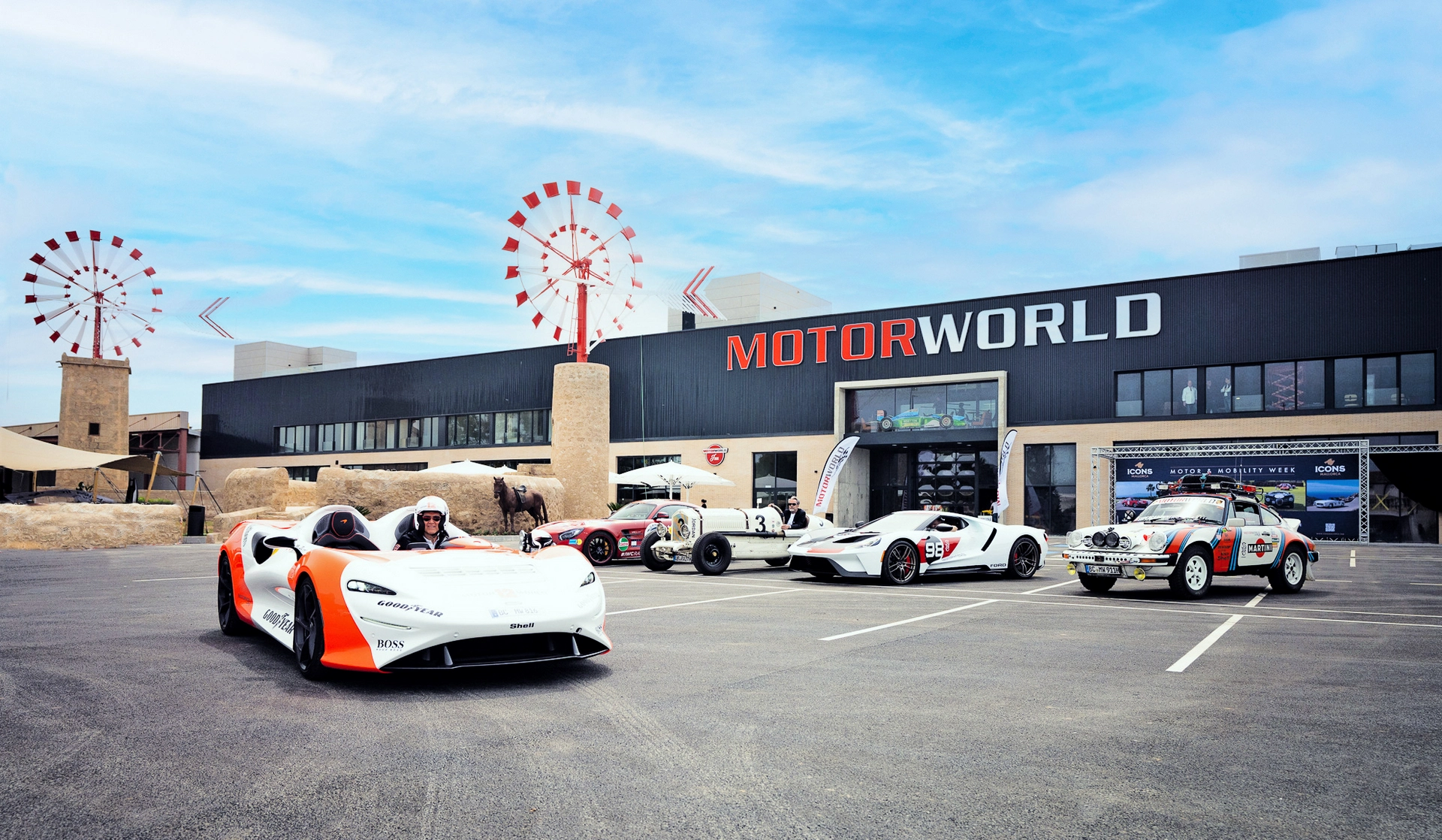 Four race cars parked in front of the MOTORWORLD building at Motorworld Mallorca, with windmills and a blue sky in the background.