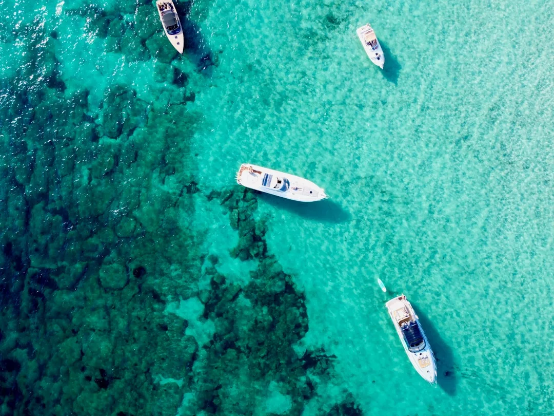 Aerial view of five boats, including a premium yacht, floating on clear turquoise water near a coral reef in Cala Ratjada.
