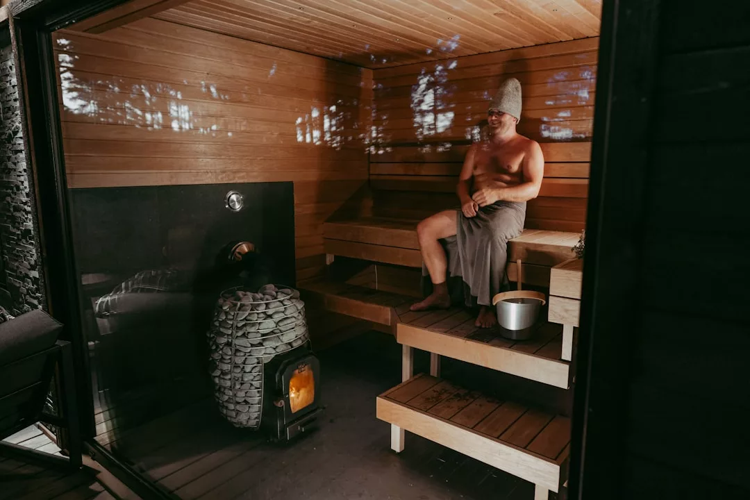A man sits on a wooden bench in a sauna, wearing a towel and hat, with a bucket beside him—enjoying exclusive wellness in one of Sant Elm's private villa saunas.