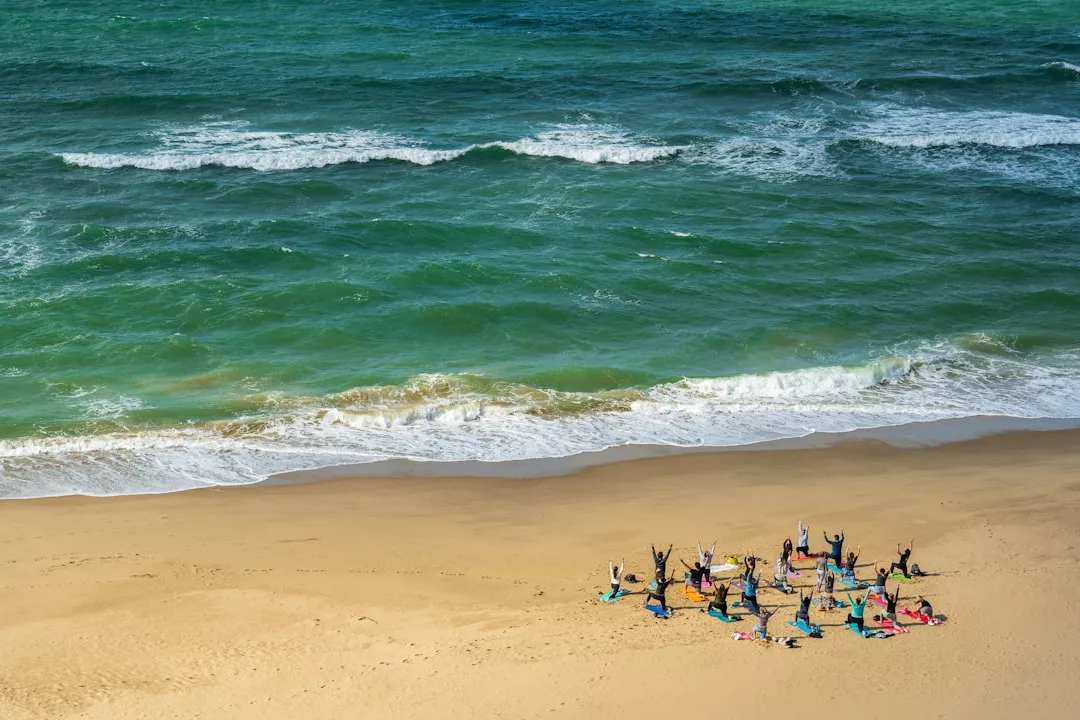 A group of people rejuvenate with yoga stretches on a sandy beach near the ocean waves under a blue sky at Colònia de Sant Jordi, perfect for wellness detox retreats.