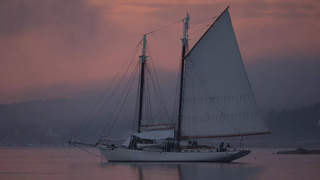 A sailboat with tall masts floats on calm water at sunset in Port d’Andratx, with a pink and purple sky in the background—perfect for scenic boat cruises.