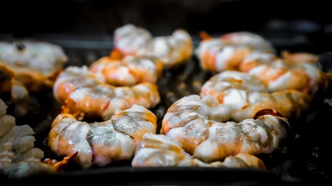 Close-up of raw shrimp, a seafood specialty often found in Port de Sóller’s beachfront restaurants, being grilled on a barbecue, with visible shells and slight charring.