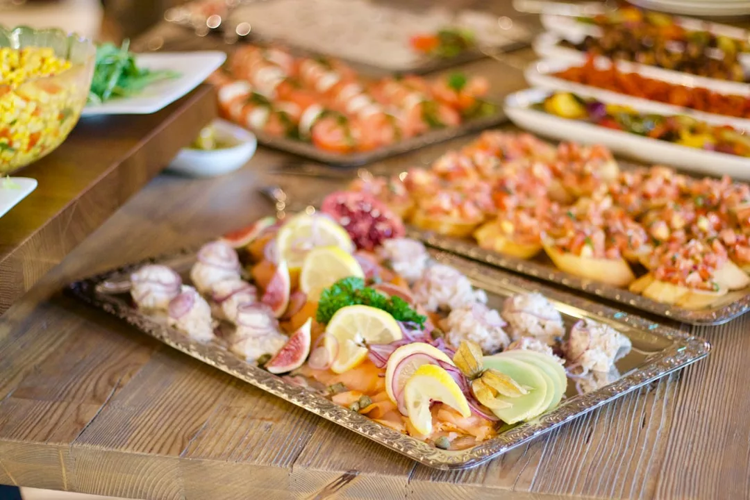 A variety of colorful appetizers and finger foods, including sustainable seafood, are arranged on platters on a wooden table.
