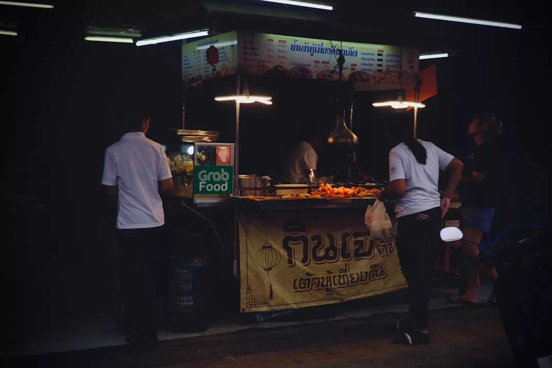Street food stall at night with people ordering food; a GrabFood sign on the counter invites you to experience an urban culinary tour reminiscent of tapas hopping in Portals Nous restaurants.