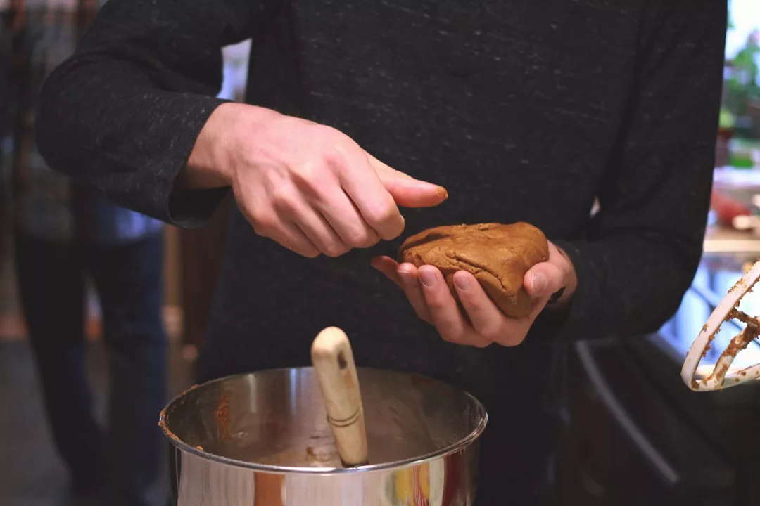 Person shaping cookie dough by hand over a mixing bowl with a wooden spoon inside, inspired by traditional Mallorcan food found in Algaida restaurants.