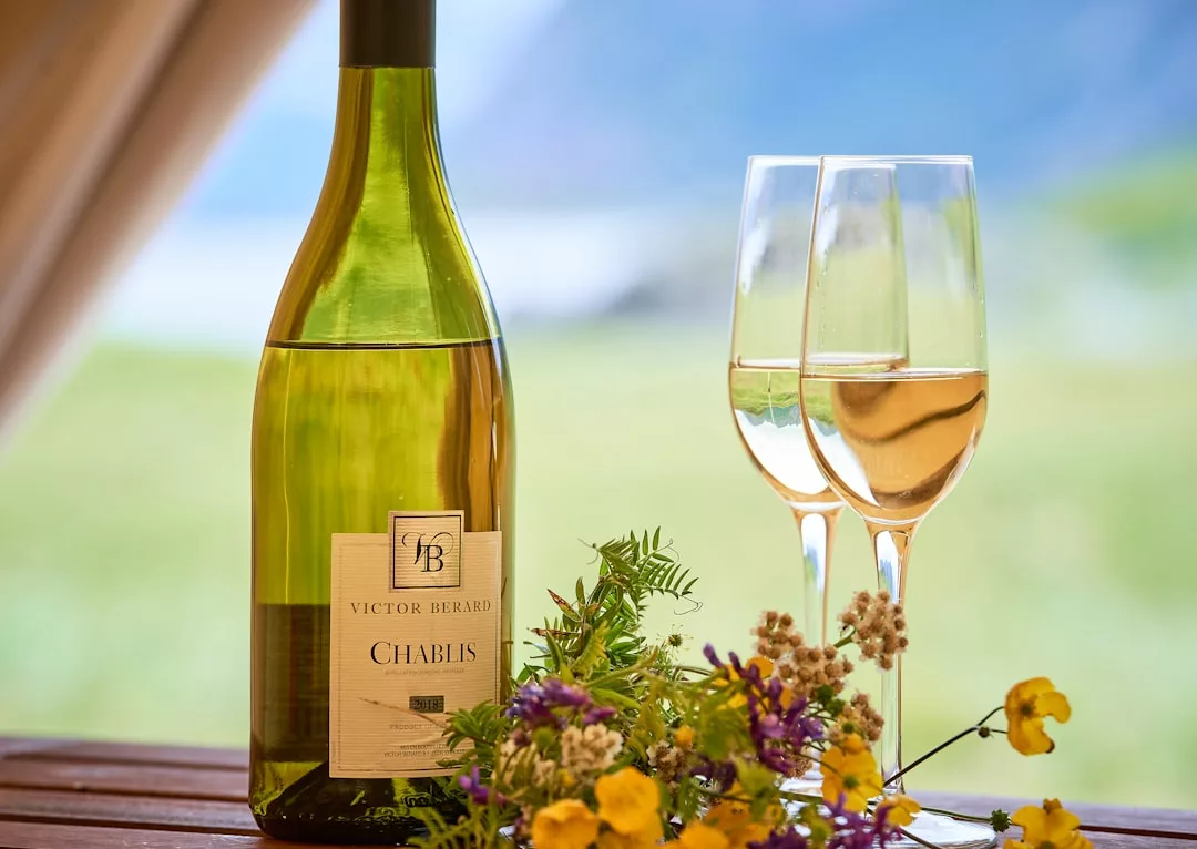 A bottle of Chablis wine and two filled glasses sit on a table with flowers outdoors, perfect for a glamping experience near Mondragó Natural Park.