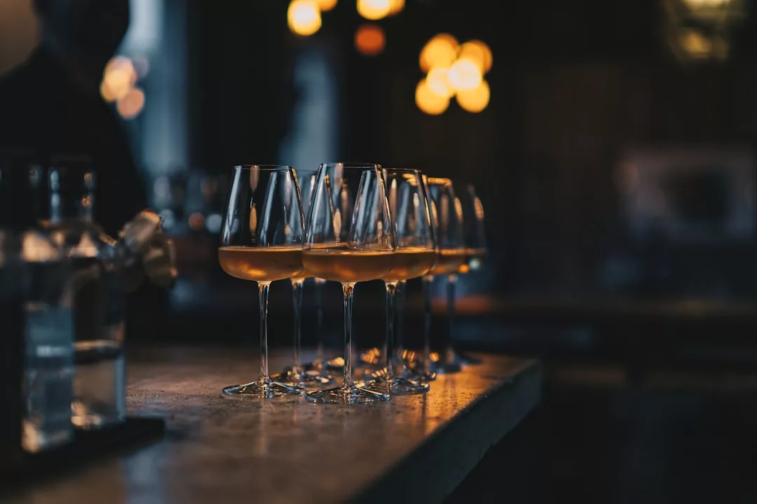 Six wine glasses filled with amber liquid sit on a dimly lit bar counter, warm lights glowing in the background—a perfect setting for wine-pairing dinners at boutique restaurants in Pollença.