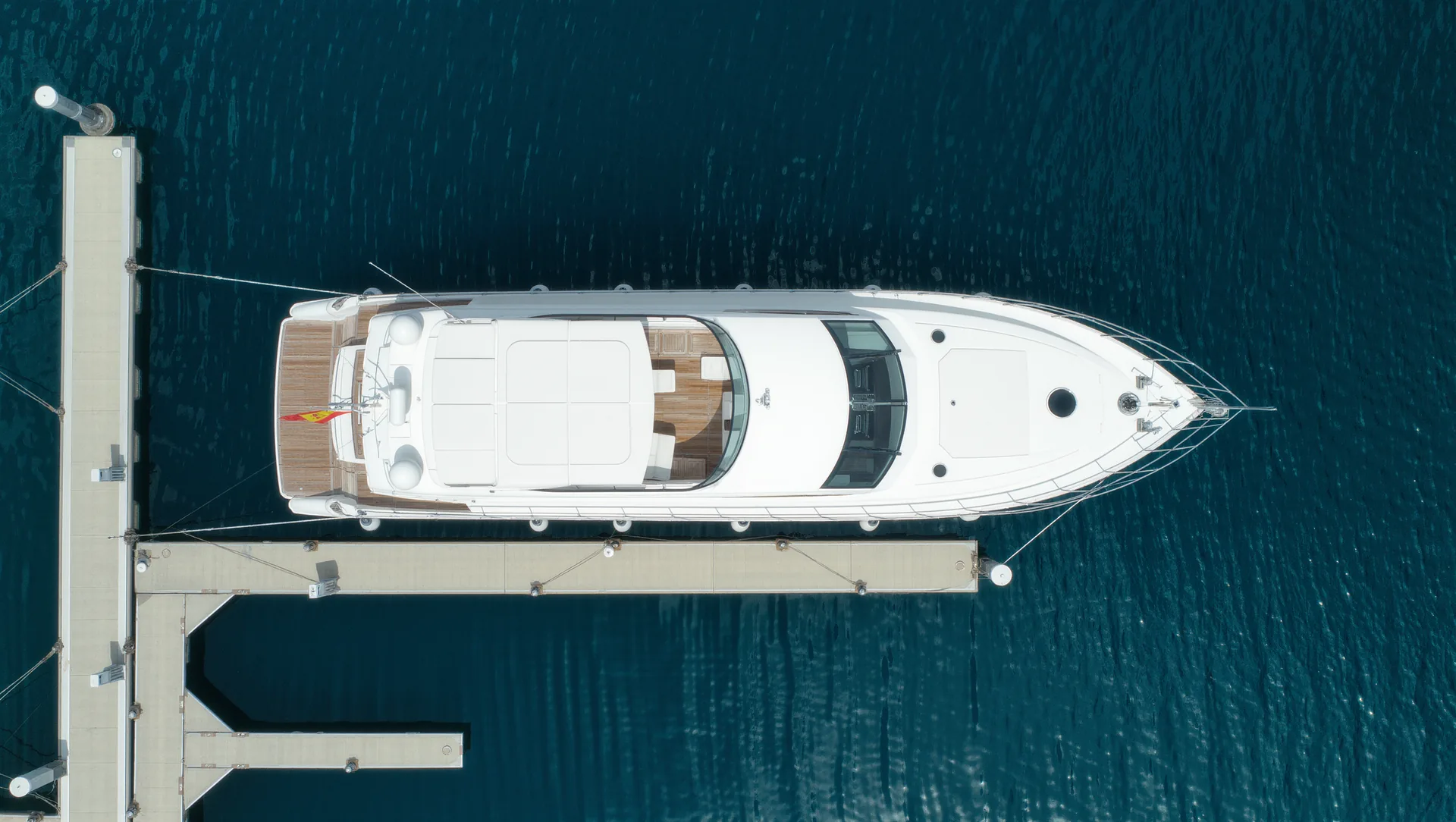 Aerial view straight down onto a white motor yacht in its berth in a Mallorcan marina with a Spanish ensign on the stern staff.
