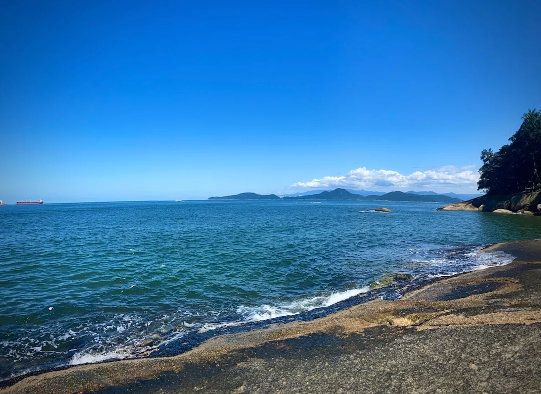 A rocky shoreline meets blue ocean waves under a clear sky, with distant mountains and a cargo ship on the horizon—an idyllic scene evoking the luxury lifestyle of Majorca.