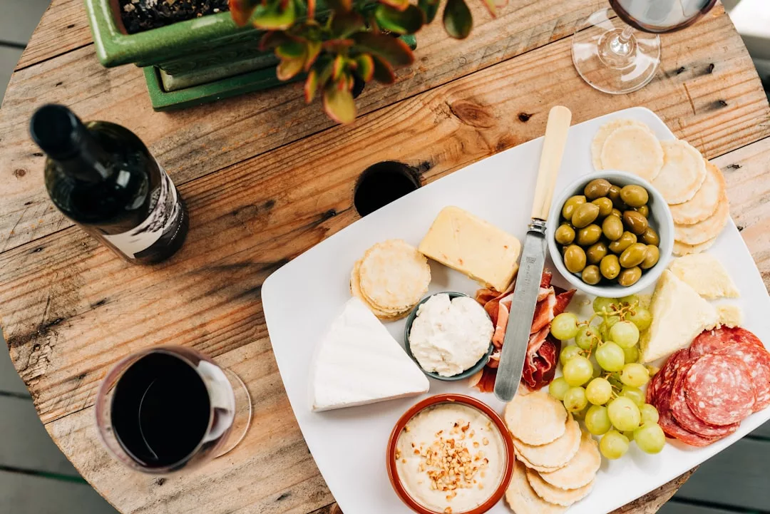 A wooden table set for a touch of luxury, with a platter of assorted cheeses, crackers, green grapes, olives, sliced meats, dips, and two glasses of red wine—perfect for savoring the lifestyle vibes of Majorca.
