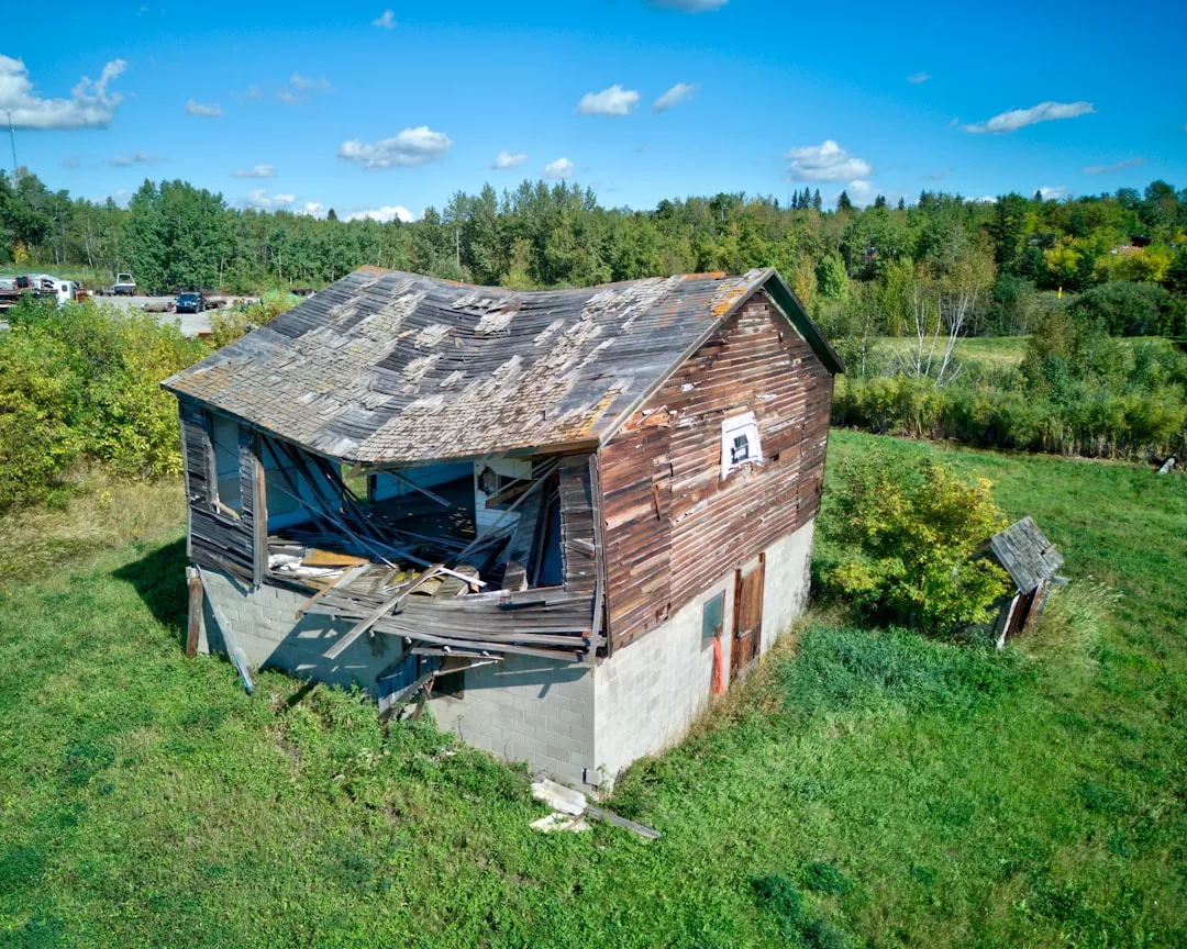 A weathered, partially collapsed wooden barn with a sagging roof stands in a grassy field, its rustic charm contrasting the luxury lifestyle often found in Majorca, surrounded by trees under a blue sky.