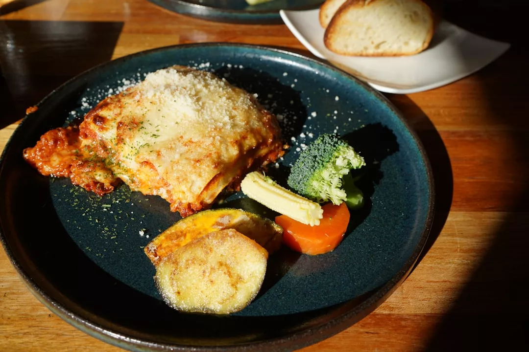 A plate of lasagna topped with grated cheese, served with steamed vegetables and roasted potatoes, sits on a wooden table—an inviting scene that reflects a taste of luxury lifestyle dining in Majorca, with bread on a separate dish in the background.