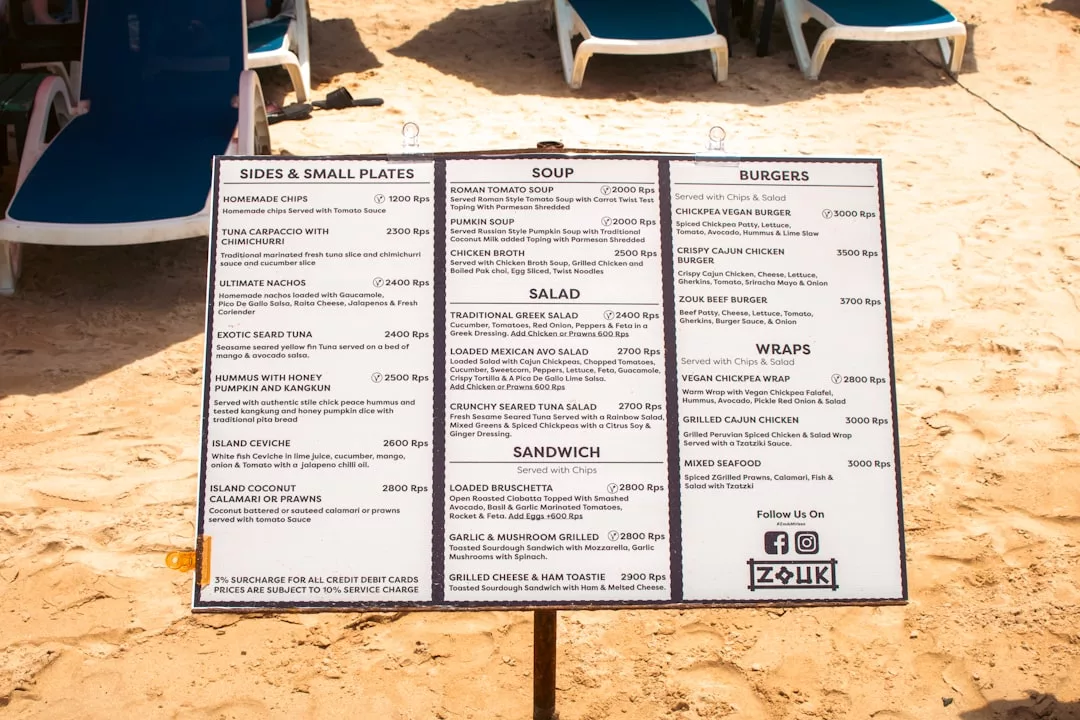 A beachside menu board on sand offers luxury sides, small plates, soups, salads, burgers, wraps, and sandwiches priced in Mexican pesos. Blue lounge chairs in the background evoke a relaxed Majorca-inspired lifestyle.