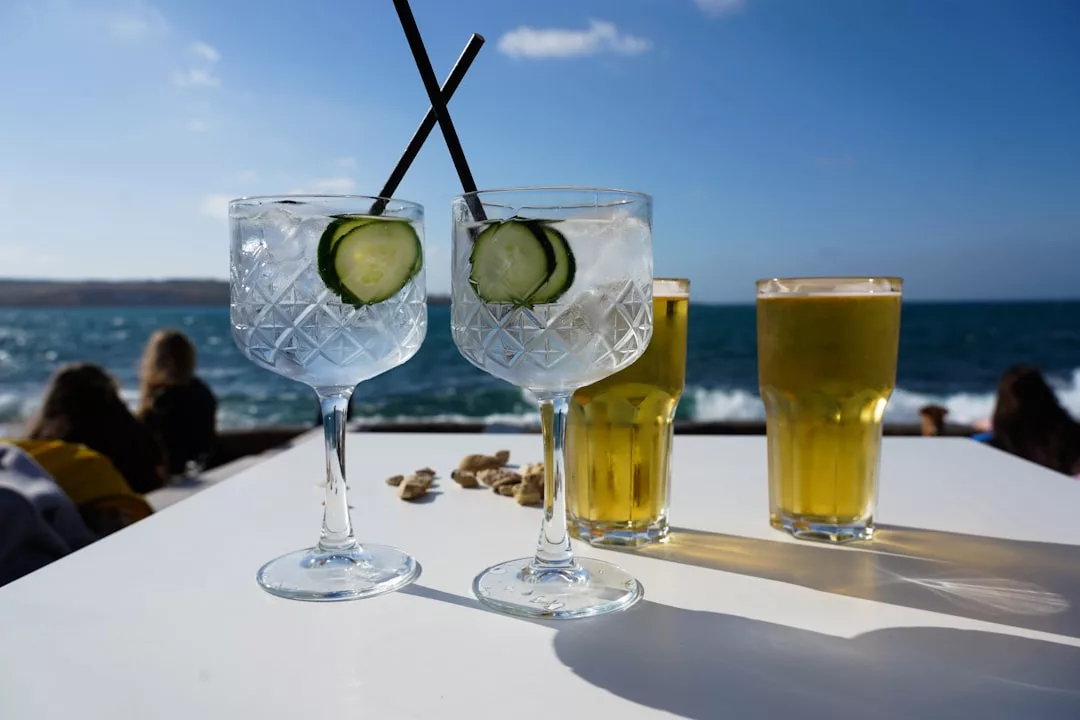 Two gin and tonics with cucumber slices and two pints of beer on a white table by the sea in Majorca, capturing a relaxed lifestyle with people and waves in the background.