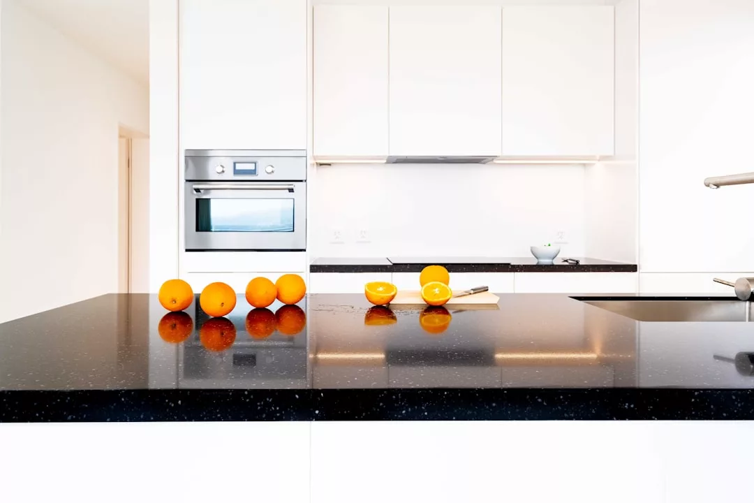 Six whole oranges and one halved orange are arranged on a black countertop, bringing a touch of Majorca luxury to this bright, modern white kitchen and elevating the lifestyle ambiance.