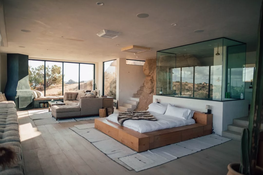 Modern luxury bedroom with glass-enclosed shower, wooden bed frame, neutral furnishings, and large windows overlooking a desert landscape, capturing the relaxed lifestyle of Majorca.
