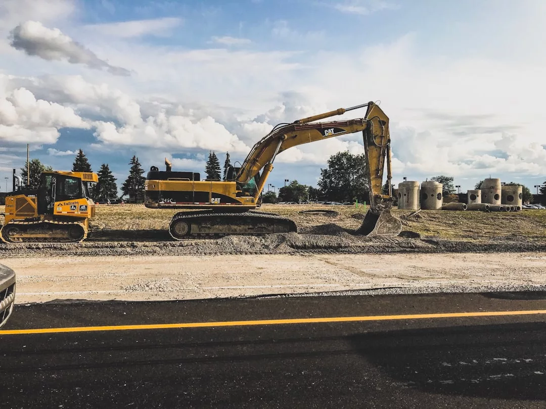 A yellow excavator scoops dirt on a construction site beside a road in Majorca, with another construction vehicle and concrete pipes nearby under a partly cloudy sky.