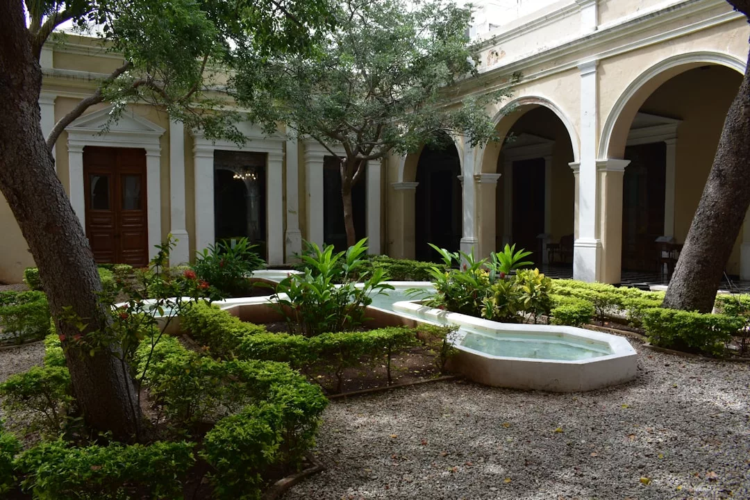 A courtyard inspired by the luxury lifestyle of Majorca, with trees, manicured bushes, and a small fountain, surrounded by arches and cream-colored walls.
